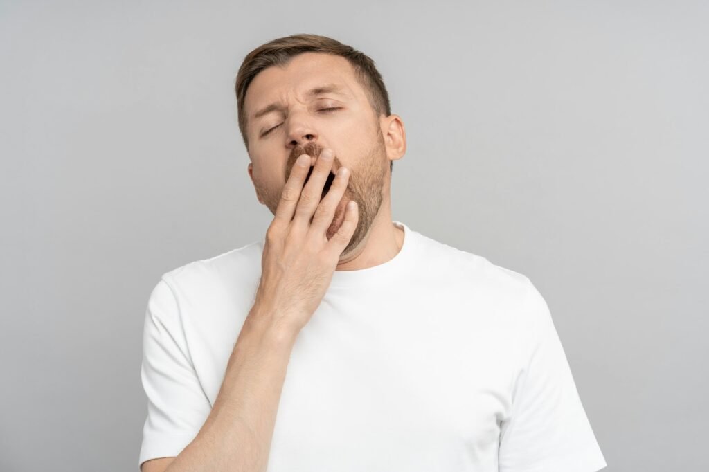 Sleepy yawning man with closed eyes opened mouth covering hand on gray background.
